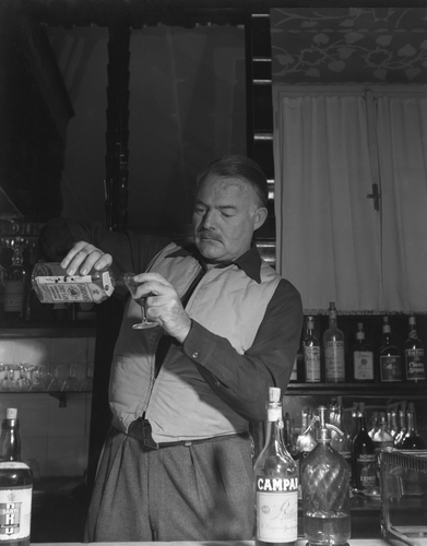 Ernest Hemingway standing behind a bar counter pouring gin from a bottle of Gordon's